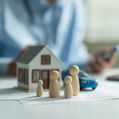 Close Up Of Wooden Family Dolls With House Model And Toy Car On Insurance Policy Document At Desk